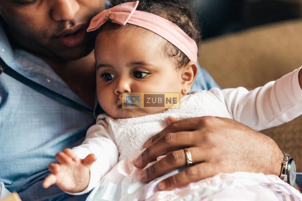 free baby images. a baby girl is sitting on couch. she is wearing a pink ribbon on her head, a white color jacket and a pink short and pink socks. her father holding her in his hand
