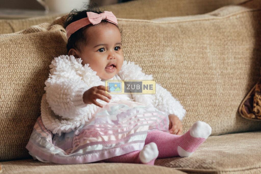 Free Baby Images. a baby girl is sitting on couch. she is wearing a pink ribbon on her head, a white color jacket and a pink short and pink socks.