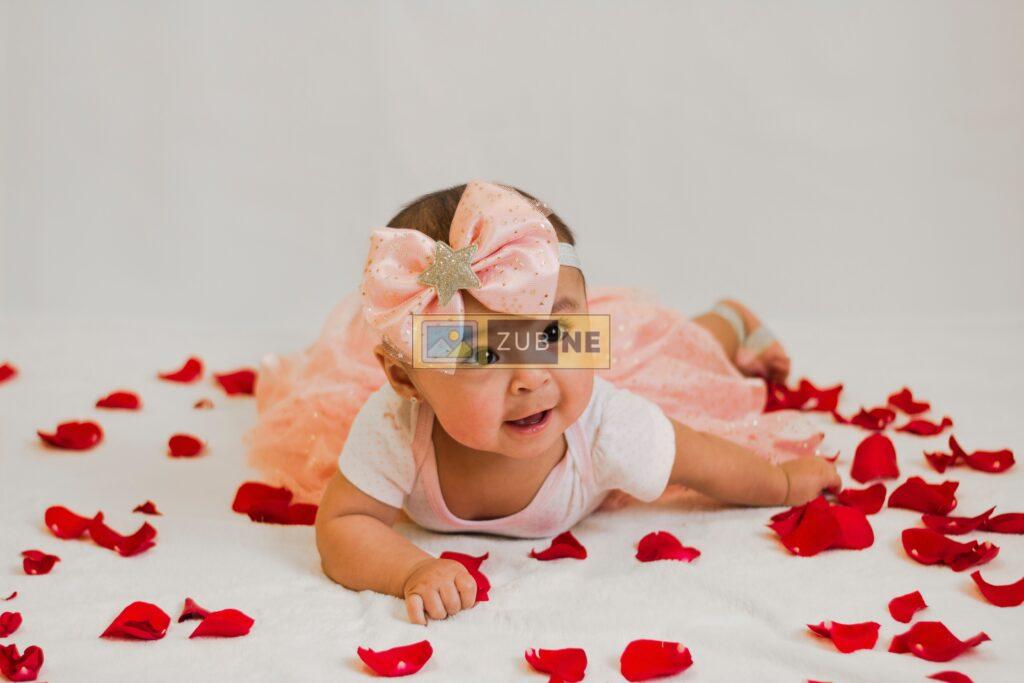A baby wearing a white dress and surrounded by rose petals
